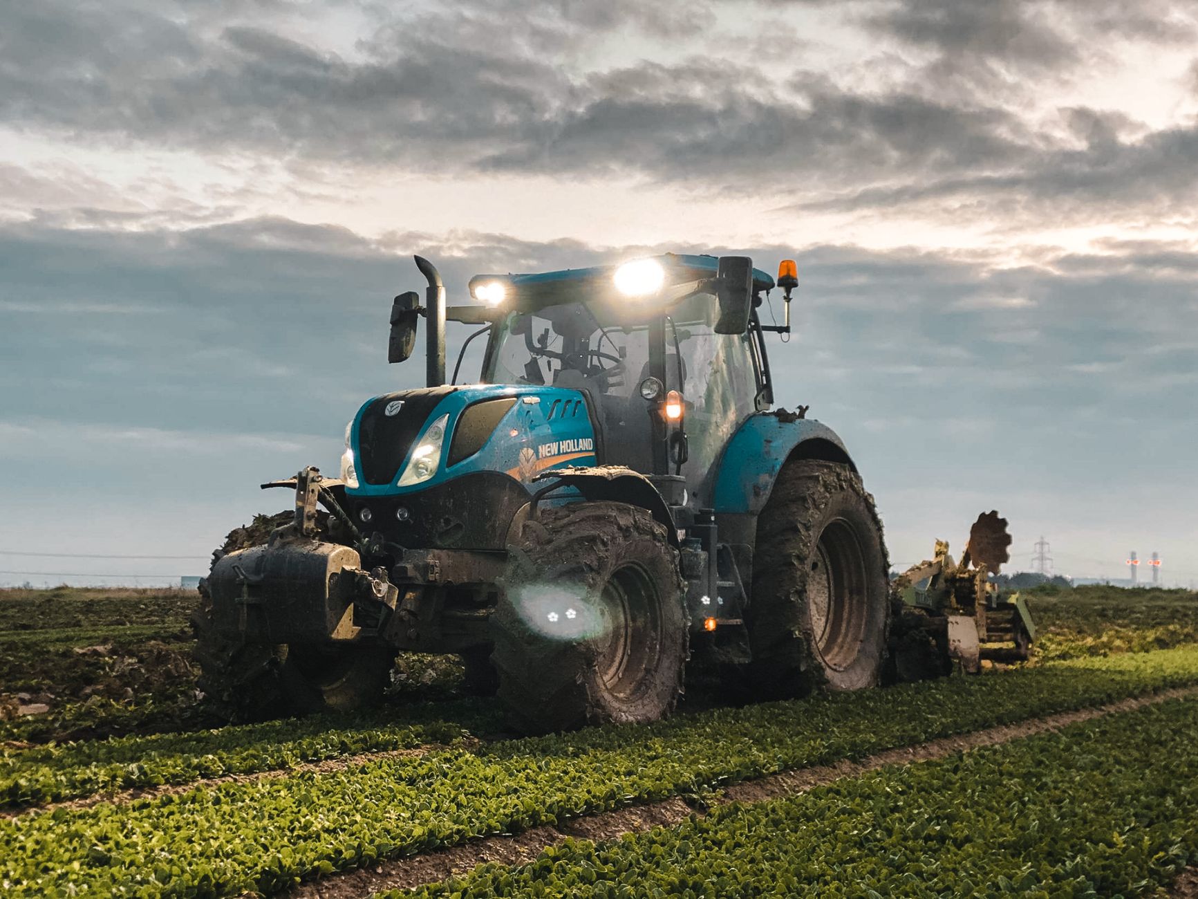 Gros tracteur avec phares et charrue traversant une plantation agricole verdoyante dans la campagne, sur des terres cultivées, sous un ciel nuageux en pleine saison des récoltes.